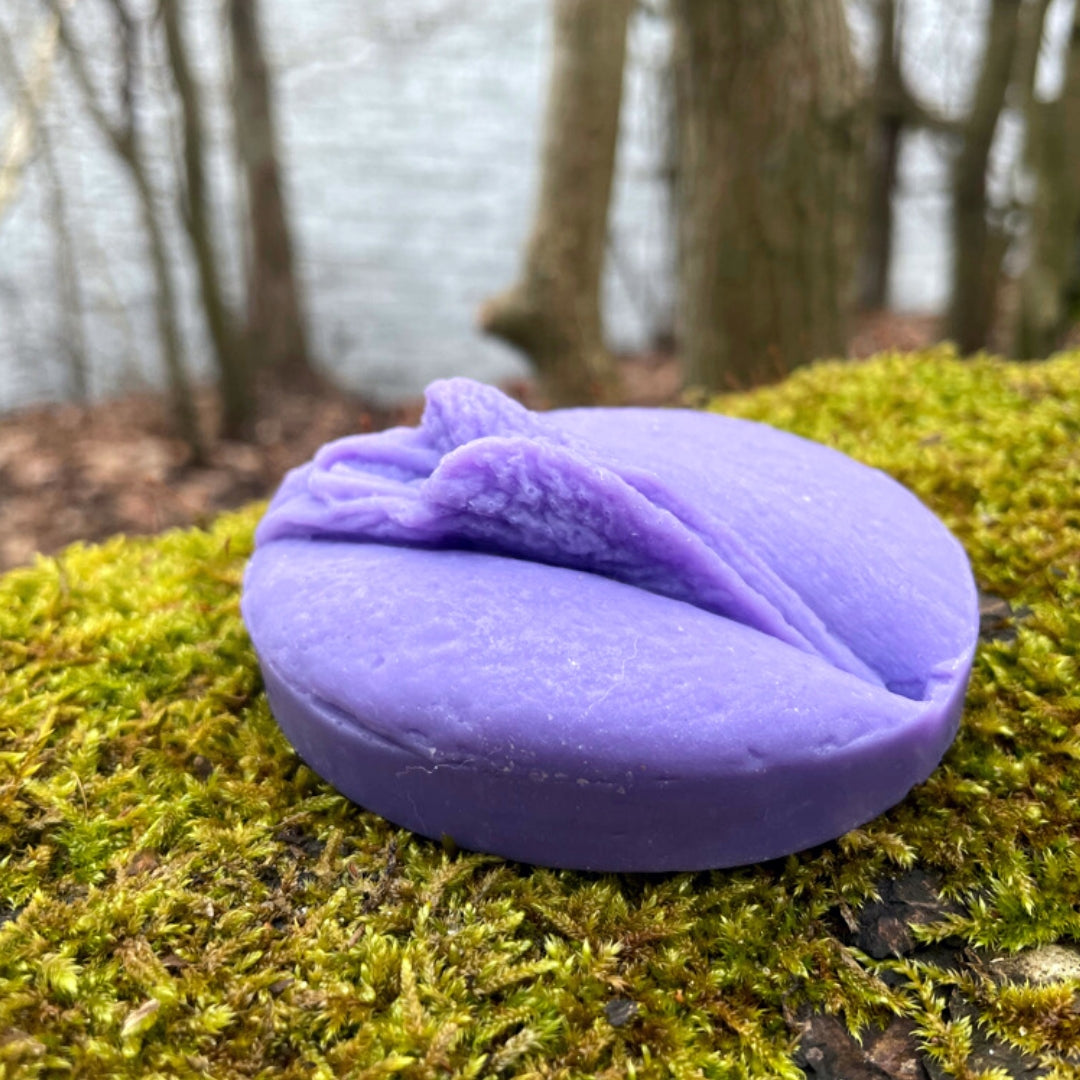 lila Vulvaseife seitlich fotografiert mit See im Hintergrund und auf Moosigem Untergrund